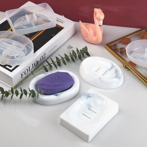 May include: Four white soap dishes with a marble pattern. Two are oval shaped and two are rectangular. One oval dish has a purple bar of soap inside. The dishes have drainage holes.