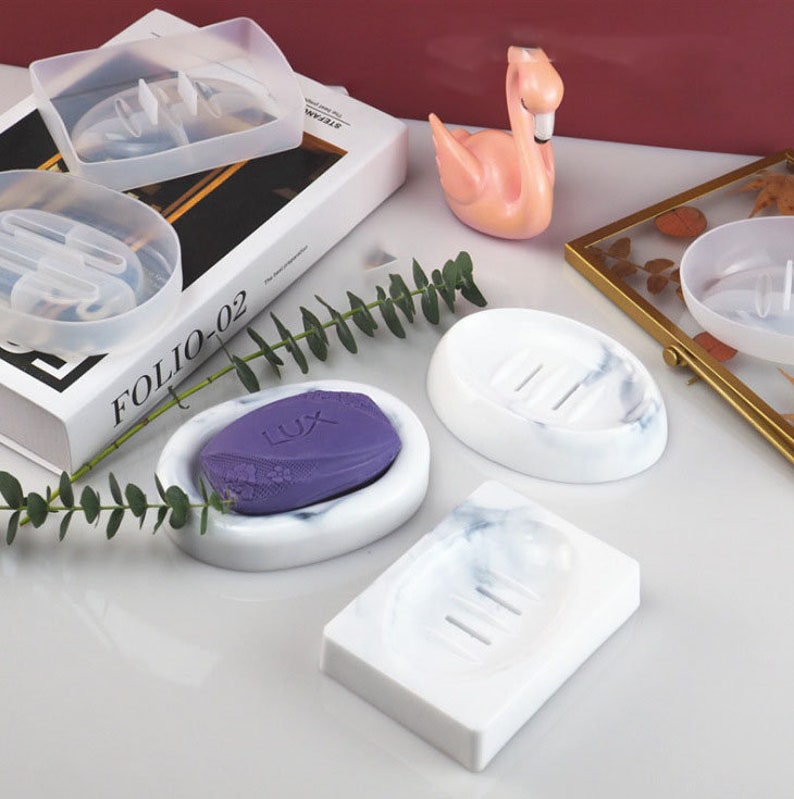 May include: Four white soap dishes with a marble pattern. Two are oval shaped and two are rectangular. One oval dish has a purple bar of soap inside. The dishes have drainage holes.