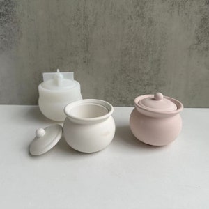 May include: Three ceramic containers with lids. One container is white, one is pink, and one is white with a lid that is off. The containers are round and have a slightly rounded top.
