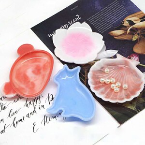 May include: Four resin trinket dishes in different shapes and colors: a pink flower, a blue cat, a red and white swirl, and a pink seashell. The dishes are on a white surface with handwritten text.