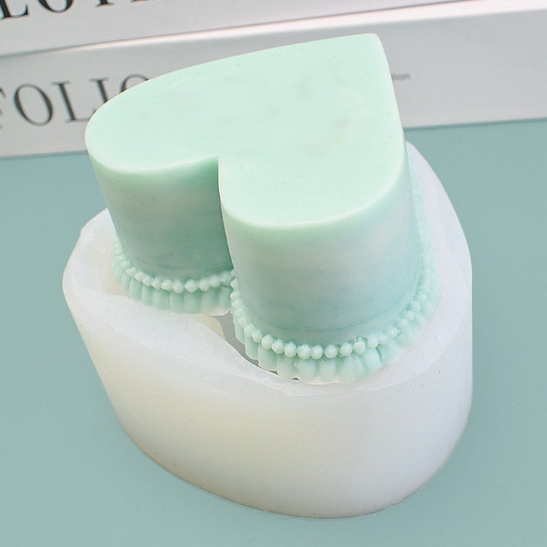 Peut inclure: Une bougie en forme de c&oelig;ur vert clair dans un moule en silicone blanc. La bougie a un motif de perles autour de la base. Le moule est de forme ovale et repose sur une surface bleu clair. La bougie est de couleur pastel.