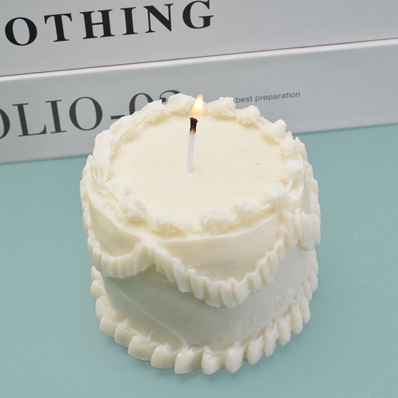 Peut inclure: Une bougie blanche en forme de g&acirc;teau avec une m&egrave;che allum&eacute;e. La bougie pr&eacute;sente un design &agrave; plusieurs niveaux avec des d&eacute;tails d&eacute;coratifs de gla&ccedil;age. Le fond comprend le mot "NOTHING" et le texte "FOLIO-03 best preparation".