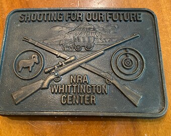 May include: A rectangular, bronze-colored belt buckle with the text "SHOOTING FOR OUR FUTURE" and "NRA WHITTINGTON CENTER." The buckle features crossed rifles, a target, a ram, and a covered wagon scene.