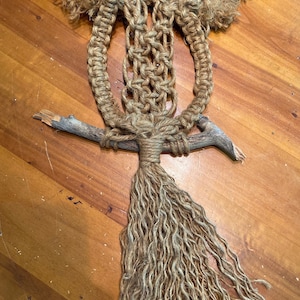 May include: Handmade macrame wall hanging featuring natural jute rope and wooden branches. The design includes a central woven pattern, a fringed bottom, and small orange beads. A rustic, bohemian style home decor item.
