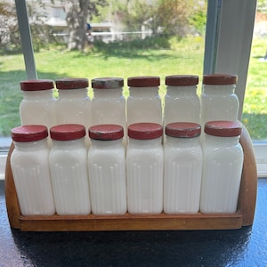 May include: A set of ten white milk glass spice jars with red lids in a wooden rack. The jars have a ribbed design and are arranged in a row.
