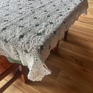 May include: Cream-colored, crocheted tablecloth with a detailed floral and geometric pattern. The tablecloth is draped over a wooden table, showcasing its intricate openwork design and scalloped edges. The table is set on a hardwood floor.