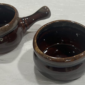 May include: Two brown ceramic soup bowls with handles. The bowls have a glossy finish and a lighter brown rim. The handles are curved and attached to the side of the bowls. The bowls are sitting on a white surface.