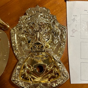 May include: A gold-toned door knocker with a lion's head design. The knocker has a crown on top and intricate details around the edges. The lion's head has a detailed face and a ring for knocking.