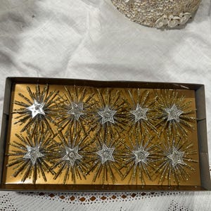 May include: A box of vintage Christmas ornaments. Each ornament is a starburst design with a silver star center and gold glittered rays. The ornaments are displayed in a gold-colored box. A decorative shell-covered item is in the background.