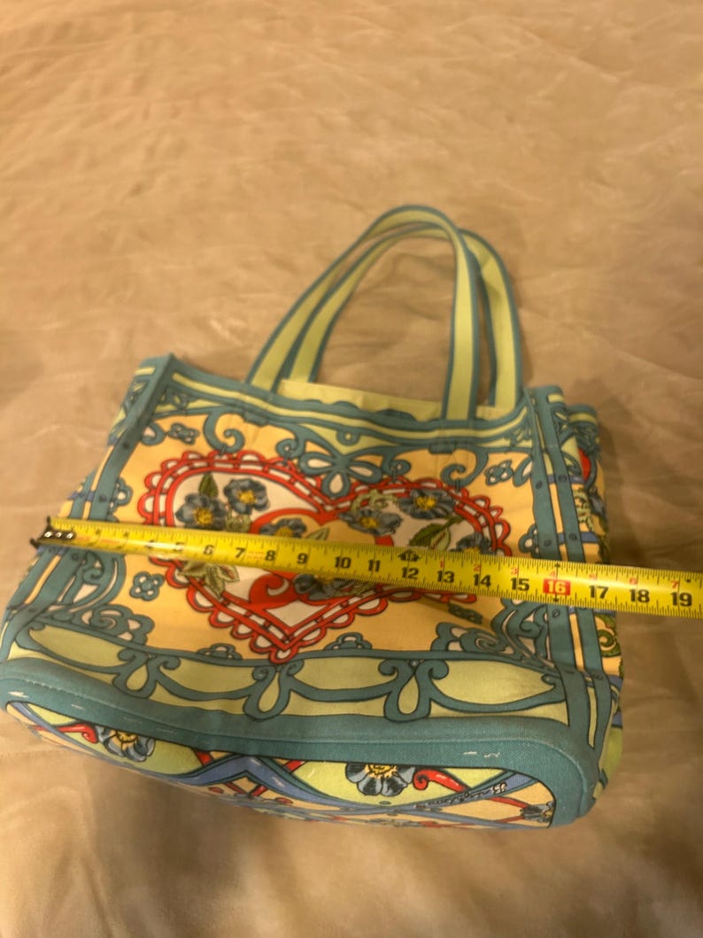 May include: A patterned tote bag with a heart design. The bag is approximately 16 inches wide. It features a yellow background with a floral and scroll design in red, blue, and green. The handles are light green.