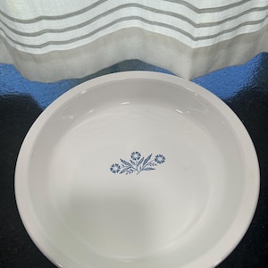 May include: White ceramic pie dish with a blue floral design in the center. The dish has a rounded shape and a slightly raised edge. The background includes a white curtain with gray stripes.