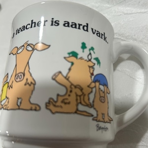 May include: White ceramic mug with a handle, featuring a cartoon aardvark design. The mug has the text "a teacher is aard vark." The aardvarks are in various poses, with green accents. The mug is a standard size for coffee or tea.