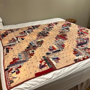 May include: A patchwork quilt with a log cabin pattern in shades of red, brown, blue, and green. The quilt is made of various fabrics and is laid out on a white bed.