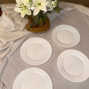 May include: Four white ceramic dinner plates on a light gray tablecloth. The plates are arranged in a circle with a bouquet of white lilies in the center.