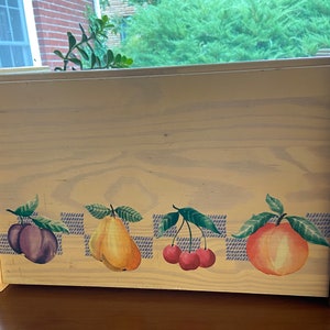May include: A wooden bread box with a light yellow finish and a painted design of fruit. The design includes a plum, pear, cherries, and a peach.