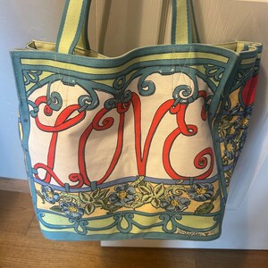 May include: Canvas tote bag with the word "LOVE" in red script. The bag features a floral and scroll design in blue, yellow, and green. The handles are striped in green and yellow.