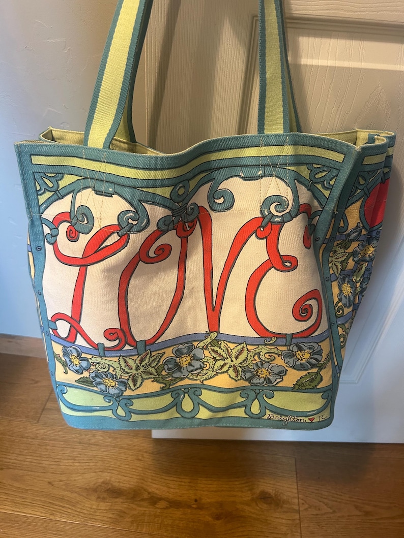 May include: Canvas tote bag with the word "LOVE" in red script. The bag features a floral and scroll design in blue, yellow, and green. The handles are striped in green and yellow.