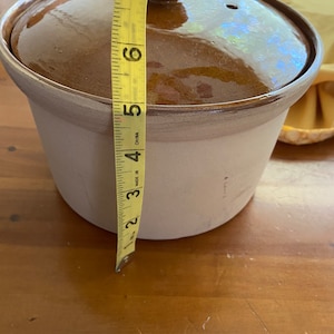 May include: A brown ceramic crock pot with a lid and a yellow measuring tape next to it. The tape reads "Made in China" and measures 7 inches.