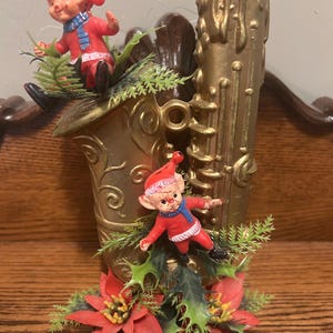 May include: A vintage Christmas decoration featuring a gold-colored saxophone with two elf figurines in red coats and hats. The saxophone is adorned with red poinsettias, green holly, and faux evergreen branches. The decoration is set on a wooden surface.