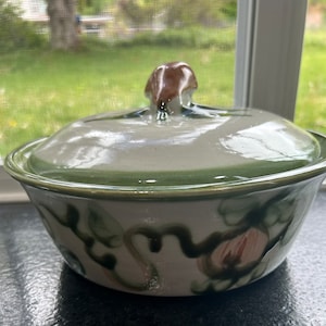 May include: A white ceramic casserole dish with a green rim and a brown knob on the lid. The dish has a floral design with green leaves and pink flowers.
