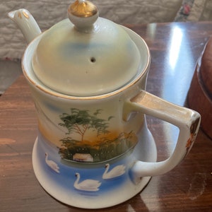 May include: A white porcelain teapot with a gold lid and handle. The teapot is decorated with a painted scene of a lake with swans and a small house in the background.