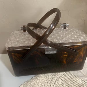 May include: A rectangular handbag featuring a dark brown tortoiseshell-patterned body and a clear, faceted, diamond-textured lid. The bag includes a single, arched, dark brown handle and a clear plastic latch closure on the front.