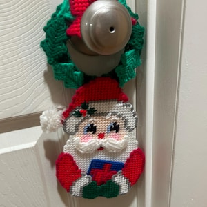 May include: A handmade Christmas ornament featuring a Santa Claus holding a gift. The ornament is made of plastic canvas and is decorated with red, white, green, and blue yarn. The ornament is hanging on a door.