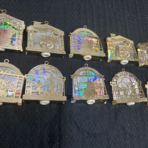 May include: A collection of gold-colored Christmas ornaments. The ornaments feature various designs, including houses and arched windows with holographic inserts. Each ornament has a small white base and a loop for hanging.