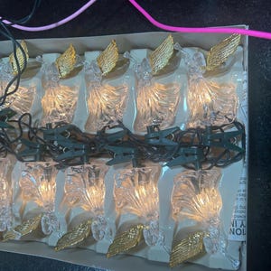 May include: A set of angel-shaped Christmas lights. Each clear plastic angel has gold-colored wings and a small light inside. The lights are strung on a green wire with clips for easy placement. The lights are packaged in a cardboard box.