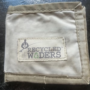 May include: A beige wallet with a stitched border. The wallet features a rectangular patch with the text "RECYCLED WADERS" and a graphic of a person fishing. The wallet is made of a durable, possibly recycled material.