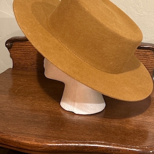 May include: A brown felt fedora hat with a wide brim. The hat is sitting on a wooden surface.