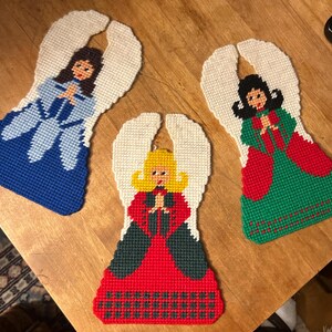 May include: Three handmade angel ornaments. Each angel has white wings and a colorful dress. One angel wears a blue dress, another a red dress, and the third a green dress. The angels are made of yarn and are likely for Christmas.