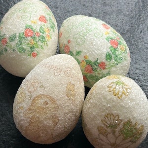 May include: Four white Easter eggs decorated with glitter and various designs. One egg has a floral pattern with red and green accents. Another egg has a floral pattern with yellow and green accents. A third egg has a floral pattern with gold accents. The fourth egg has the text "Happy Easter" written on it.