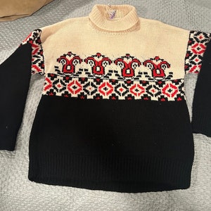 May include: A black and cream turtleneck sweater with a red and black geometric pattern. The sweater has a traditional Scandinavian design.