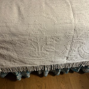 May include: A textured, off-white bedspread with a fringed edge. The bedspread covers a bed skirt with a blue and white floral pattern. The bed is positioned over a hardwood floor.