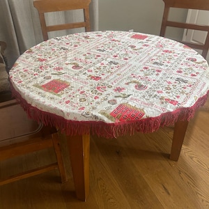 May include: A round tablecloth with a white background featuring a repeating pattern of red houses, birds, flowers, and the text "HOME SWEET HOME". The fabric is finished with a decorative red fringe trim along the entire circular edge.