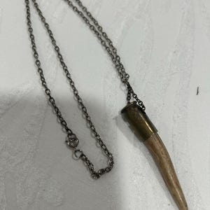 May include: A necklace featuring a pendant made from a light brown antler, capped with a brass-colored metal. The pendant hangs from a delicate, dark silver-toned chain. The necklace is displayed on a white surface.