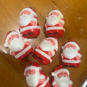 May include: A collection of seven small Santa Claus figurines. Each figurine is red and white, with a white beard, a red hat, and a black base. The figurines are arranged on a wooden surface.