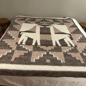 May include: A white and brown alpaca wool throw blanket with a geometric pattern featuring two llamas. The blanket is on a bed with a white bedspread.
