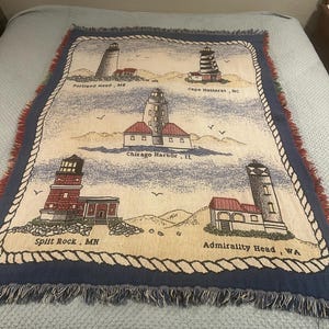 May include: A woven throw blanket featuring five lighthouses with the text Portland Head, ME; Cape Hatteras, NC; Chicago Harbor, IL; Split Rock, MN; and Admiralty Head, WA. The design is framed by a rope border on a blue background with fringe edges.