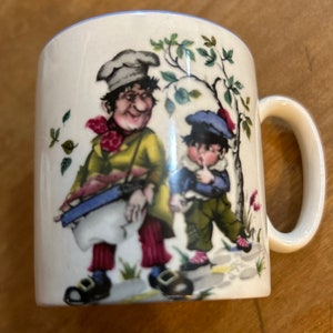 May include: White ceramic mug with a whimsical illustration of two figures in a garden setting. One figure wears a chef's hat and carries a drum. The other figure is a young boy. The mug has a light blue rim and a curved handle.