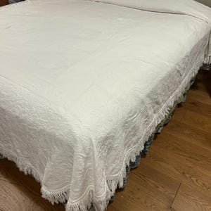 May include: A white bedspread with a detailed floral pattern covers a bed. The bedspread has a fringe trim along the edges. The bed is on a hardwood floor. A small wooden nightstand is visible in the background.