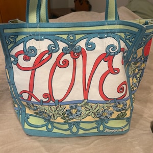 May include: A canvas tote bag with a white background and the word "LOVE" in red script. The bag features a floral design with blue flowers and green leaves, and has blue and green trim. The handles are striped.