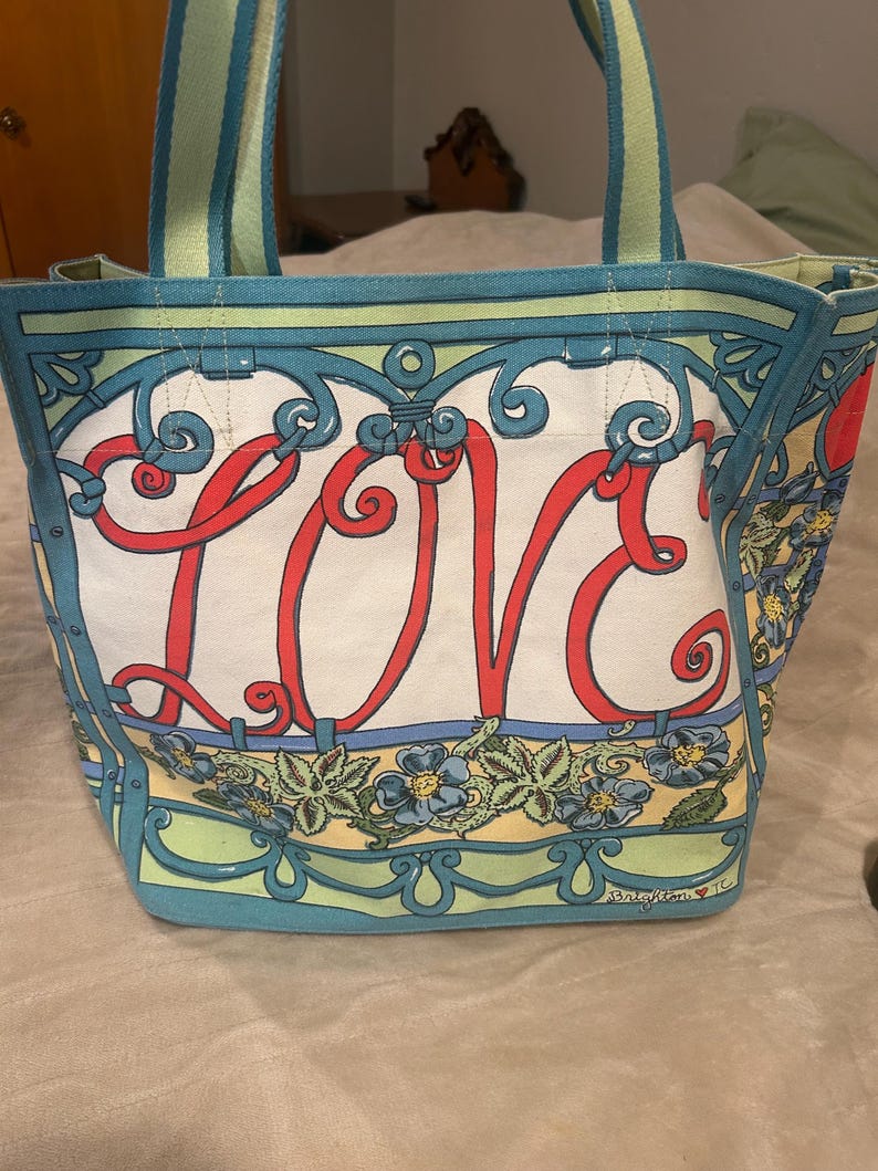 May include: A canvas tote bag with a white background and the word "LOVE" in red script. The bag features a floral design with blue flowers and green leaves, and has blue and green trim. The handles are striped.