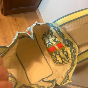 May include: Open view of a patterned tote bag with a matching zippered pouch. The bag features a yellow background with blue and red decorative elements. The interior is a light beige color, and the straps are yellow with a green trim.