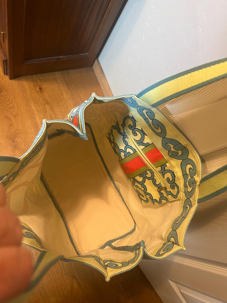 May include: Open view of a patterned tote bag with a matching zippered pouch. The bag features a yellow background with blue and red decorative elements. The interior is a light beige color, and the straps are yellow with a green trim.