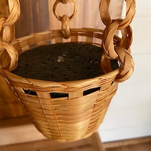 May include: A hanging planter made of woven straw with a dark gray liner. The planter is suspended by a chain made of woven straw.