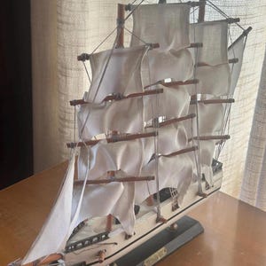 May include: A detailed model of a sailing ship with white sails and wooden masts. The ship is painted white with a black hull and sits on a dark gray base. The sails are billowing, suggesting the ship is at sea.