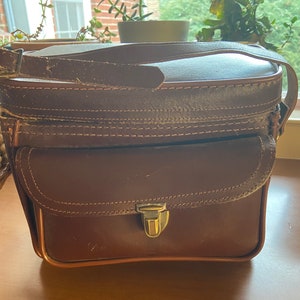 May include: A brown leather camera bag with a flap closure and a brass buckle. The bag has a front pocket and a strap.