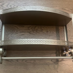 May include: A gold-toned metal wall shelf featuring two tiers with arched, decorative lattice-patterned railings. The unit includes a horizontal bar at the bottom supported by scrolled metal accents, providing a functional storage structure.
